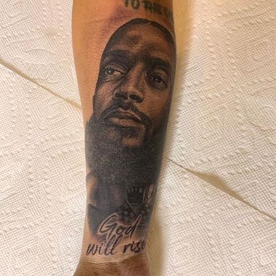 The tattoo has Nipsey Hussle's face with the text that says God will rise right below it.